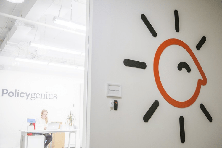 the Policygenius office with its logo mark on the wall of the entrance. The receptionist sits at their desk working in the background.