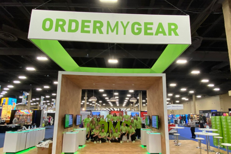The OrderMyGear display at a tradeshow. Screens showing different information are displayed inside a wooden cube while member of the team stand around the OMG Logo.